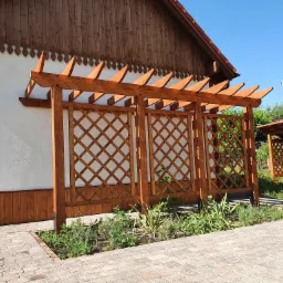 Drewniana pergola z kratownicą, przylegająca do białej ściany budynku z drewnianą podbitką, otoczona kostką brukową i rabatą z roślinami w Wąbrzeźnie. W tle widoczna kolejna, podobna konstrukcja.