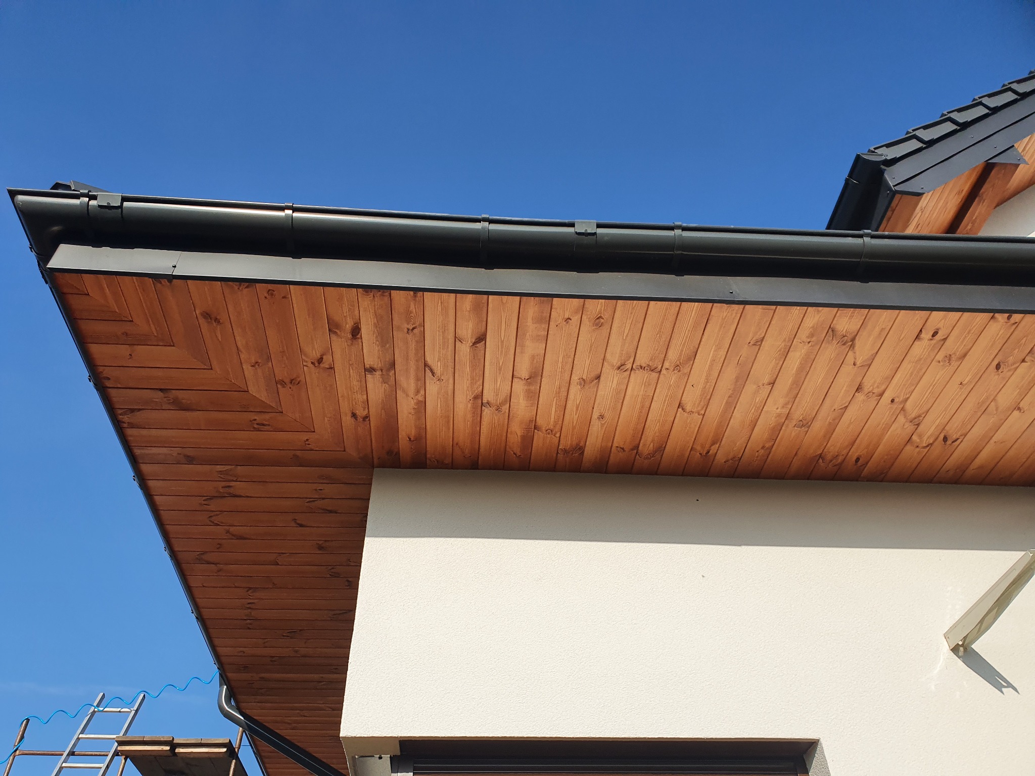 Drewniana podbitka dachowa z czarną rynną, widok z dołu na tle błękitnego nieba, drabina oparta o ścianę budynku.