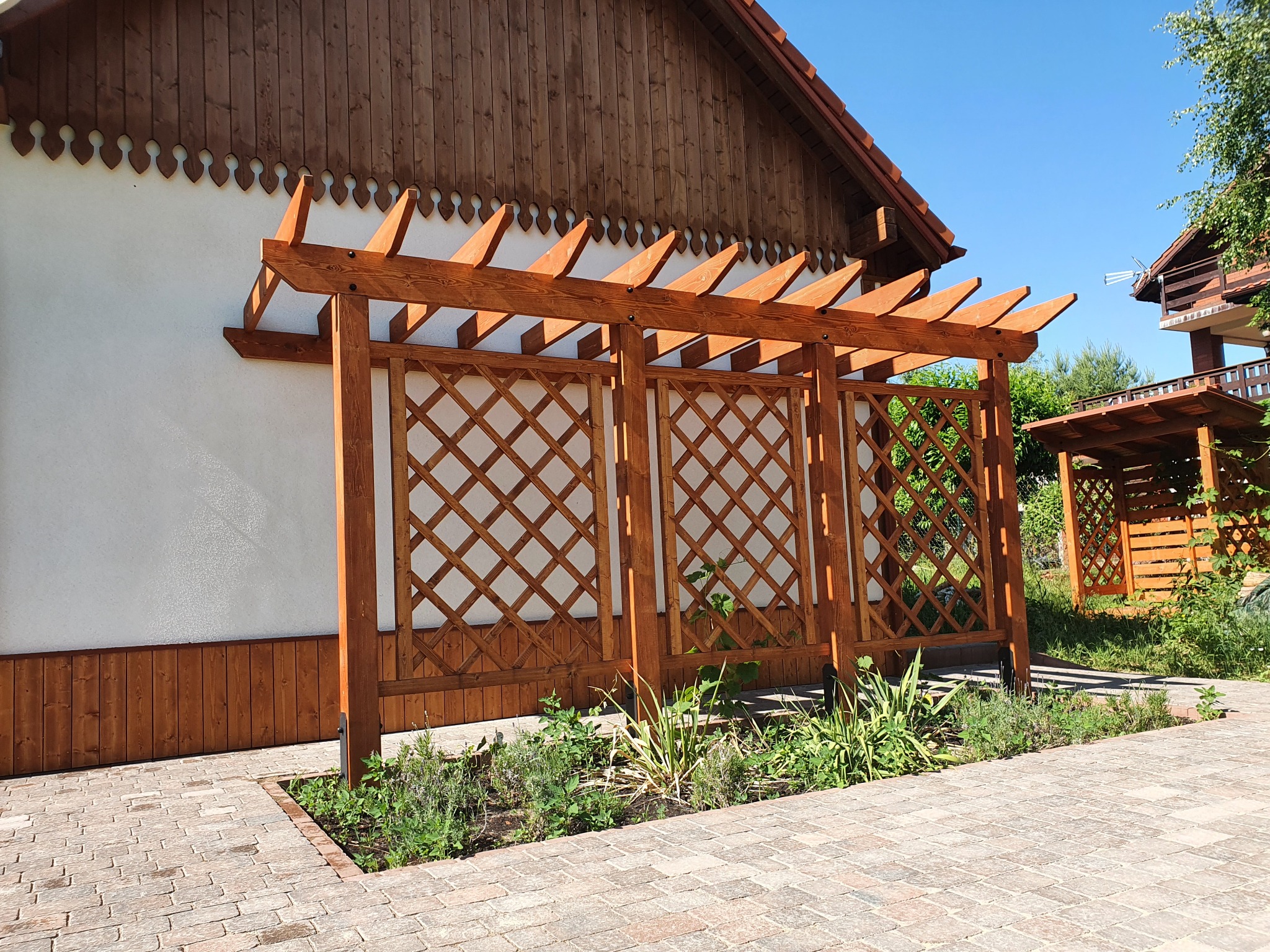 Drewniana pergola z kratownicą, przylegająca do białej ściany budynku z drewnianą podbitką, otoczona kostką brukową i rabatą z roślinami w Wąbrzeźnie. W tle widoczna kolejna, podobna konstrukcja.