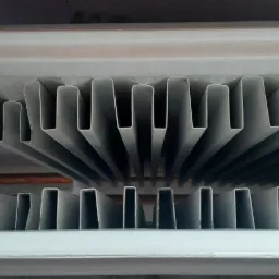 Boczny widok na żeliwny grzejnik żeberkowy, z widocznymi pionowymi żebrami i rurami doprowadzającymi wodę, wymagający czyszczenia.