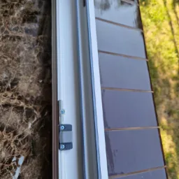 Otwarty fragment białej ramy okiennej z widocznym mechanizmem blokującym, z brązowym marmurowym parapetem wewnętrznym i błyszczącym, brązowym parapetem zewnętrznym z widocznym trawnikiem w tle.