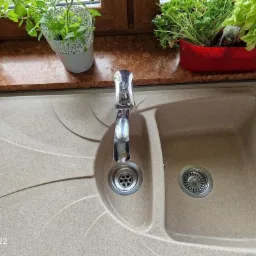 Beżowy, dwukomorowy zlewozmywak granitowy z chromowaną baterią, na tle okna z roślinami doniczkowymi.