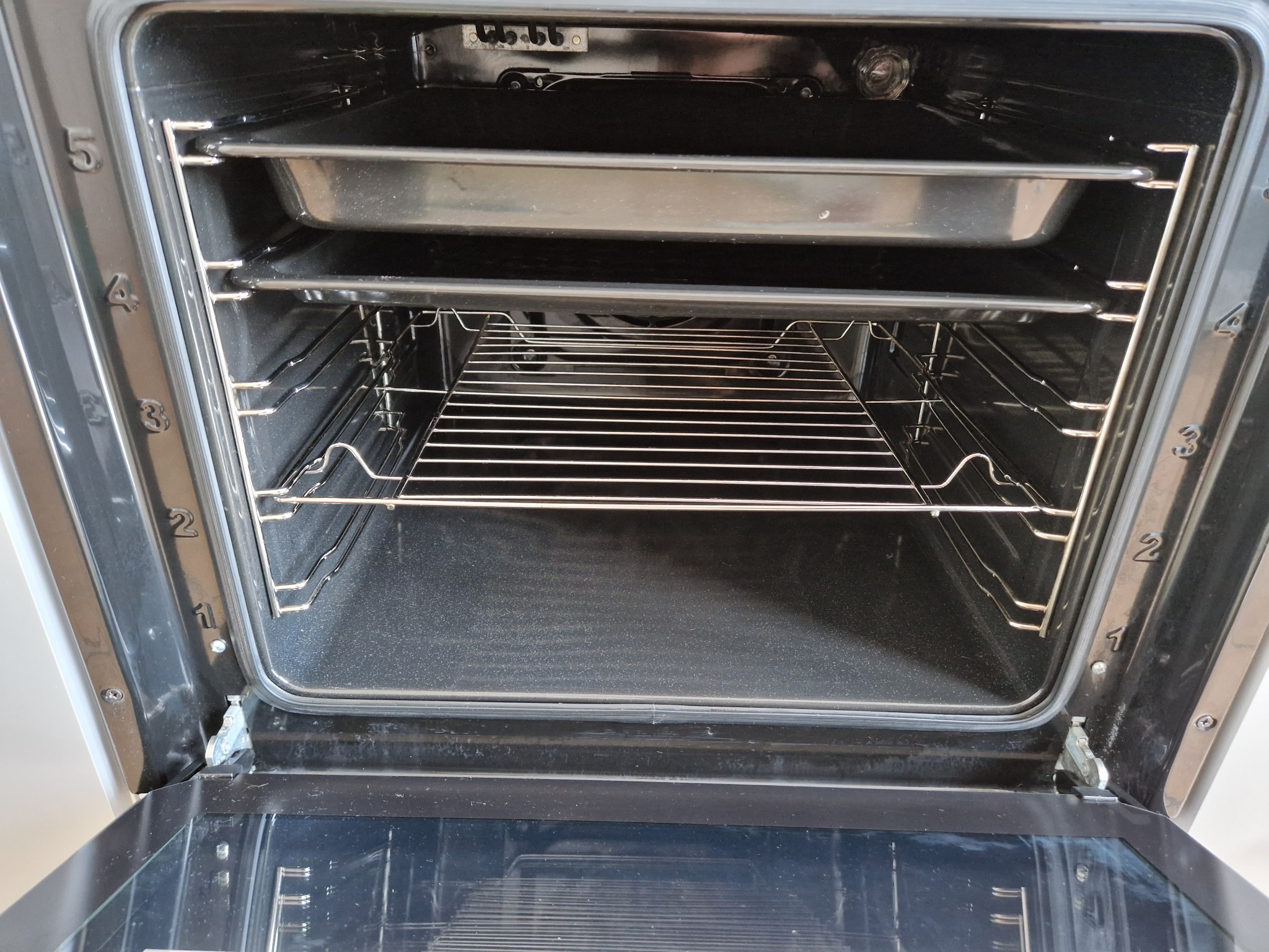 Wnętrze czystego piekarnika elektrycznego z metalową kratką i blachą, widoczne numery poziomów na ściankach bocznych.