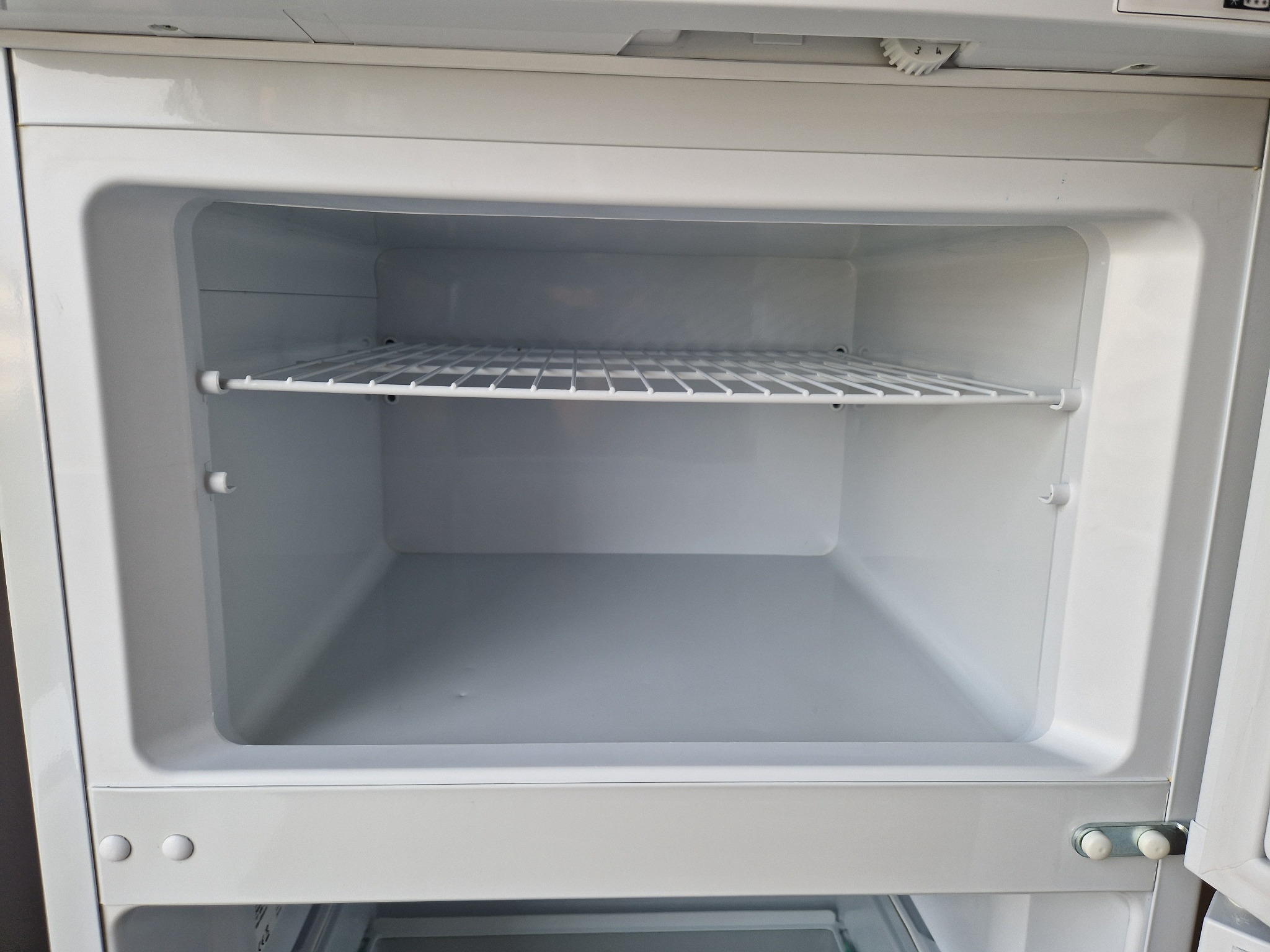 Wnętrze czystej, pustej, białej lodówki z jedną metalową półką kratową, widoczne elementy zawiasów drzwi.