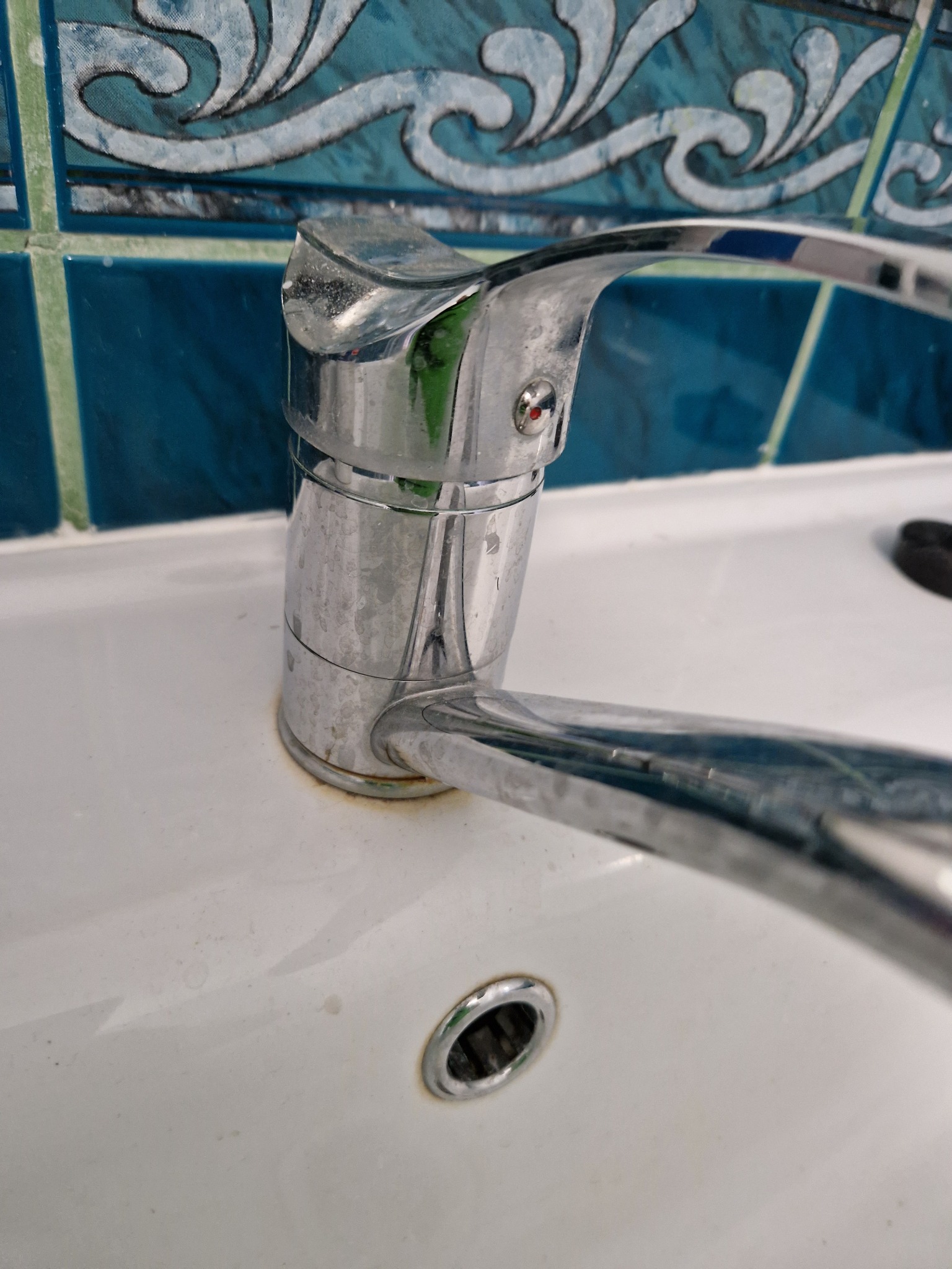 Chromowana bateria umywalkowa z widocznymi śladami kamienia i zaschniętych kropli na tle niebieskich płytek z wzorem fal. Umywalka z drobnymi zabrudzeniami.