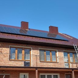 OLE PV Fotowoltaika - Częściowo zamontowane panele słoneczne na brązowym, blaszanym dachu dwupiętrowego domu z czerwonej cegły, drabina oparta o dach, błękitne niebo w tle.