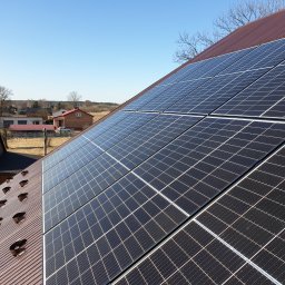 OLE PV Fotowoltaika - Panele słoneczne na brązowym, blaszanym dachu domu jednorodzinnego, widoczne elementy mocujące, w tle zabudowa wiejska i błękitne niebo.
