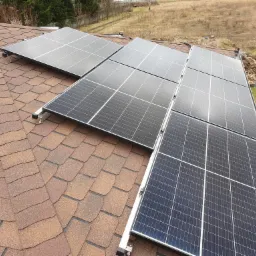 Panele słoneczne zamontowane na dachu pokrytym brązową dachówką bitumiczną, widok z góry, z widocznymi elementami mocującymi i otoczeniem budynku.