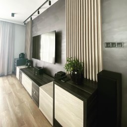 FIX HOUSE Kamil Dziedzic - Nowoczesny salon z szarą ścianą, drewnianymi lamelami dekoracyjnymi, podświetleniem szynowym, niską szafką RTV z białymi frontami i drewnianą podłogą.