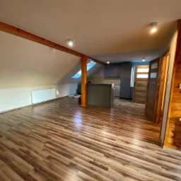 Przestronne poddasze z drewnianą konstrukcją stropu, wykończone panelami podłogowymi w odcieniach brązu, z widoczną kuchnią w tle i grzejnikiem przy drewnianej ścianie.