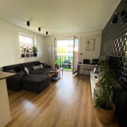 FIX HOUSE Kamil Dziedzic - Jasne wnętrze salonu z drewnianą podłogą, czarną narożną sofą, otwartym balkonem i ścianą wyłożoną błyszczącymi, czarnymi płytkami.