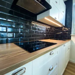 Nowoczesna kuchnia z czarnymi, błyszczącymi płytkami typu metro, drewnianym blatem, białymi szafkami z chromowanymi uchwytami i płytą indukcyjną.