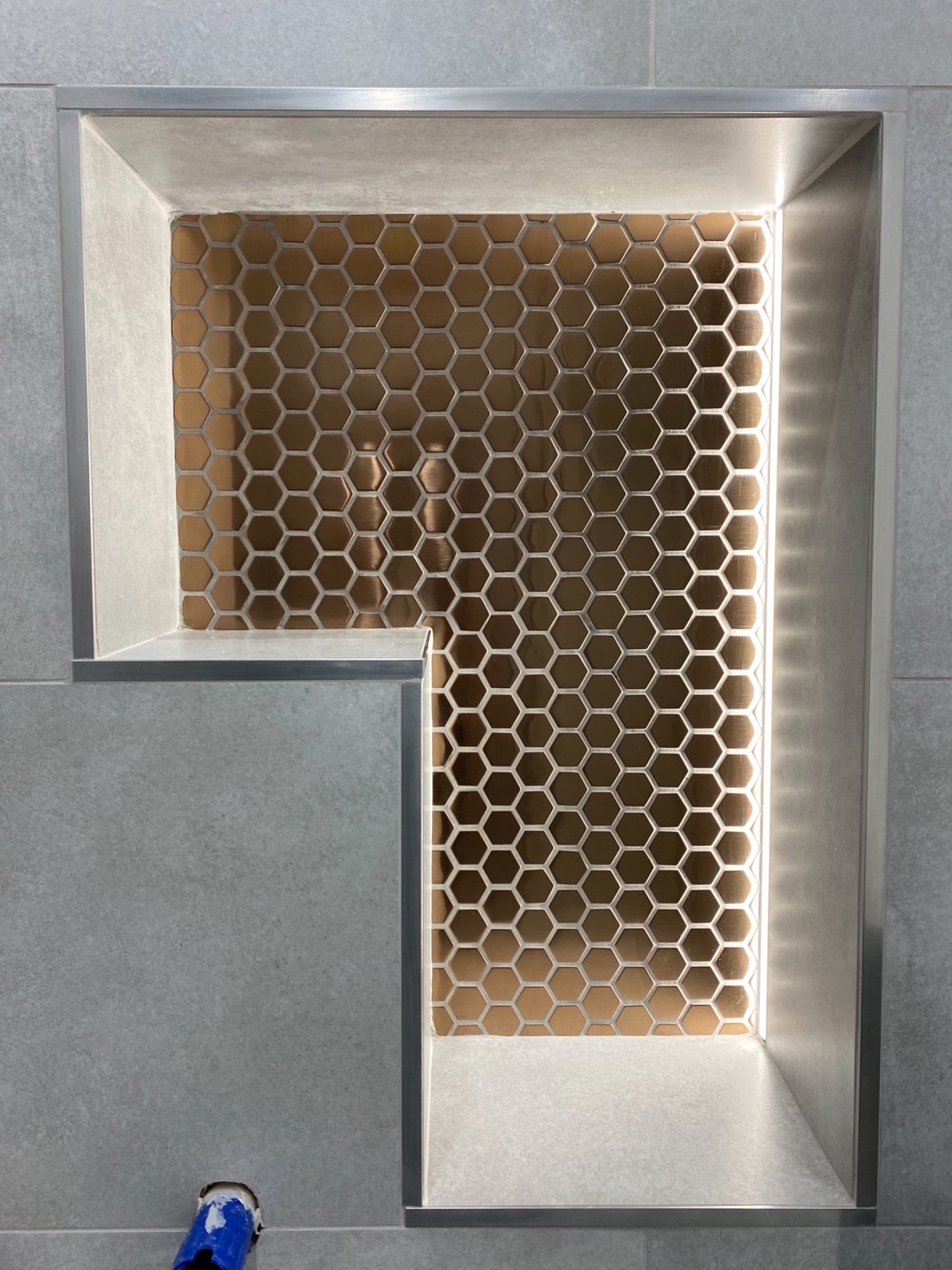 Wnęka w łazience wyłożona błyszczącą, miedzianą mozaiką heksagonalną z podświetleniem LED, otoczona szarymi płytkami i metalowymi listwami wykończeniowymi.