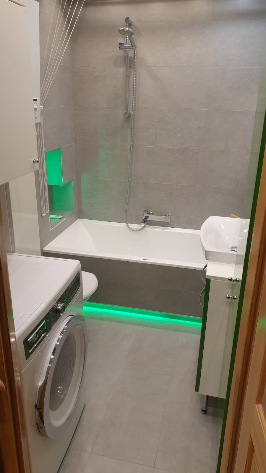 Nowoczesna łazienka z szarymi płytkami, oświetleniem LED w kolorze zielonym pod wanną i w półkach ściennych, pralką i umywalką z szafką.