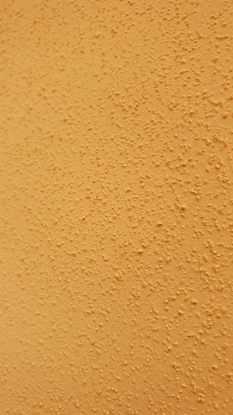 Pomarańczowa ściana z wyraźną, nierównomierną strukturą, przypominającą baranek lub ziarno, oświetlona z boku, tworząca delikatny cień.