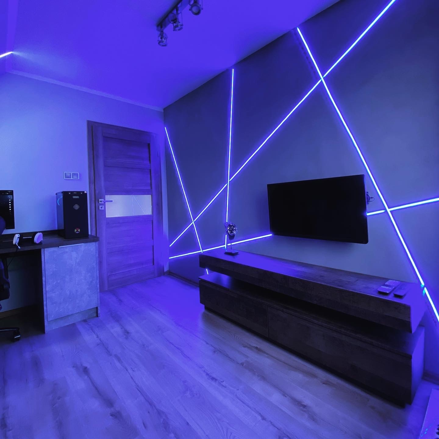 Pokój z drewnianą podłogą i ścianą zdobioną neonowymi liniami w kolorze niebieskim, oświetlony niebieskim światłem, z telewizorem na ścianie i biurkiem z komputerem po lewej stronie.