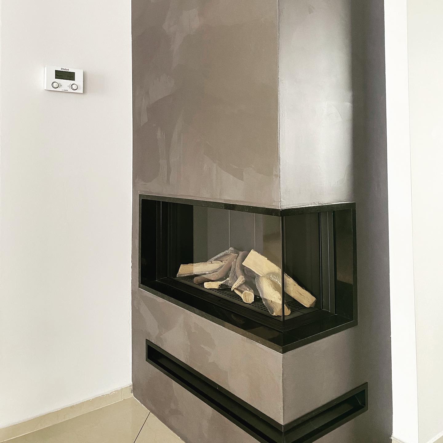 Narożny kominek z szybą, obudowany szarym tynkiem dekoracyjnym o nieregularnej fakturze. W tle biała ściana z zamontowanym panelem sterowania ogrzewaniem.