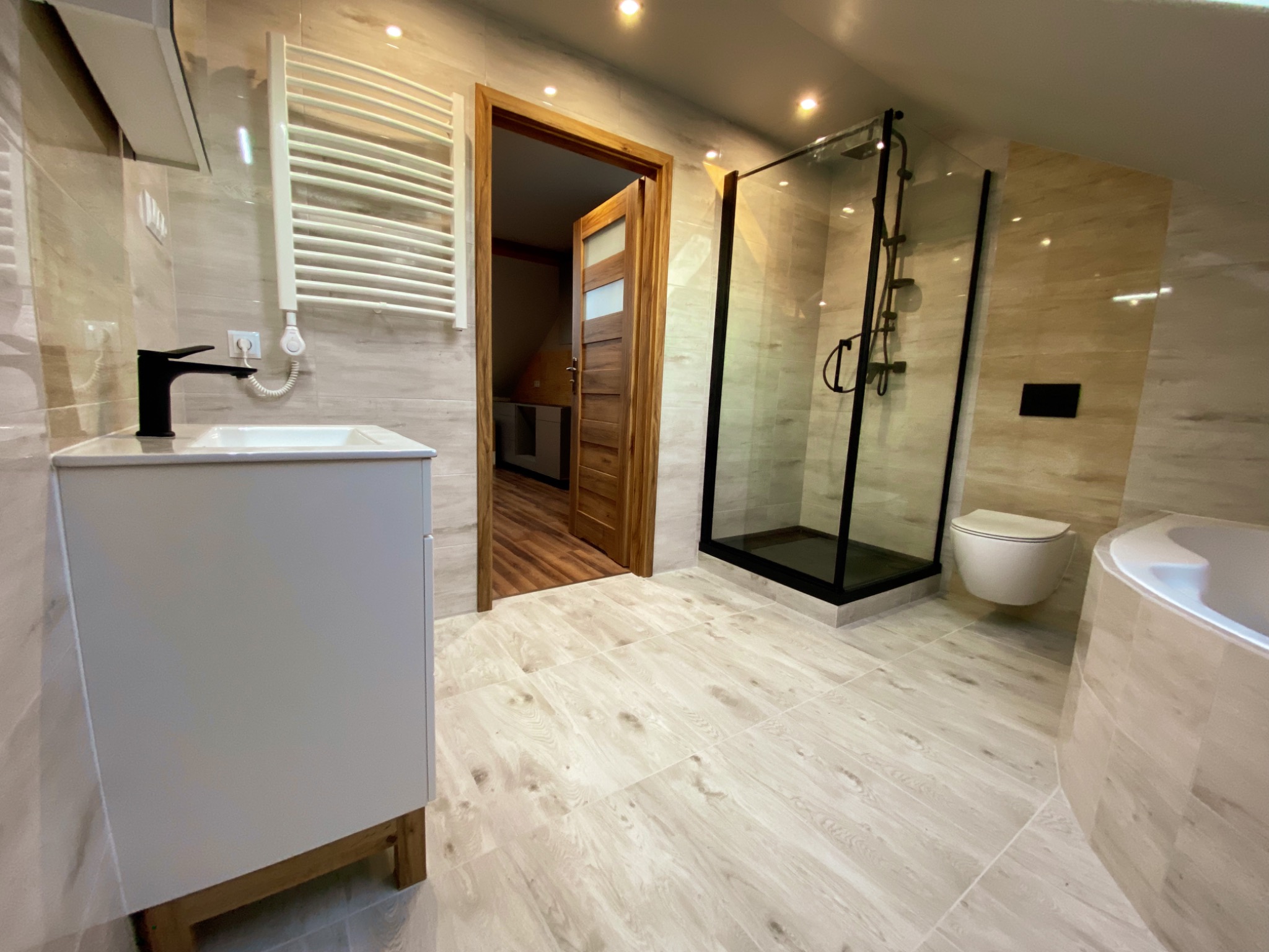 Nowoczesna łazienka na poddaszu z beżowymi płytkami imitującymi drewno, kabiną prysznicową z czarnymi ramami, wiszącą toaletą, białą umywalką z czarną baterią i drewnianymi drzwiami z przeszkleniami.