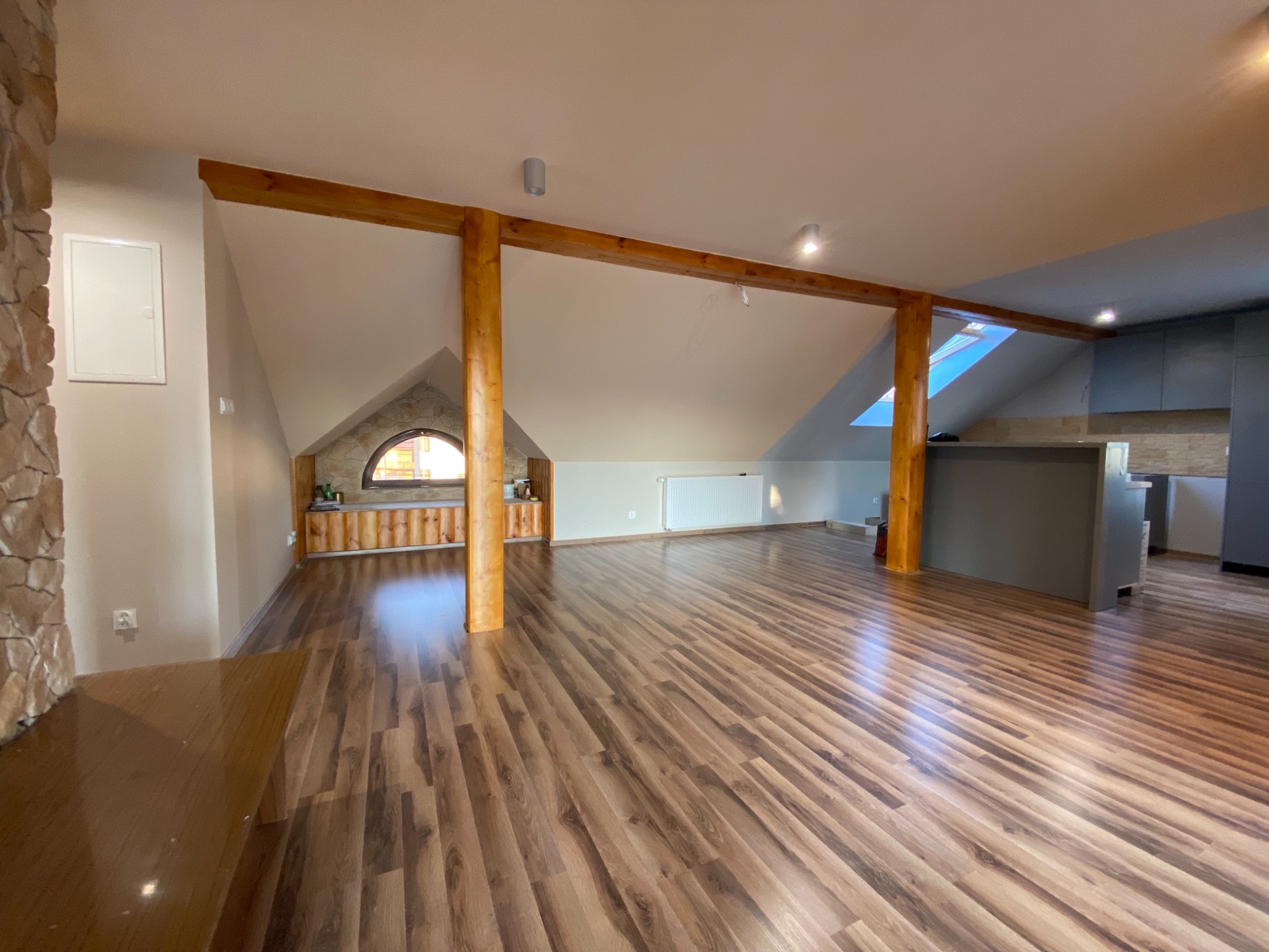 Przestronne poddasze z drewnianą podłogą w odcieniach brązu, widoczne drewniane belki konstrukcyjne, okno w kształcie łuku i fragment zabudowy kuchennej w szarościach.