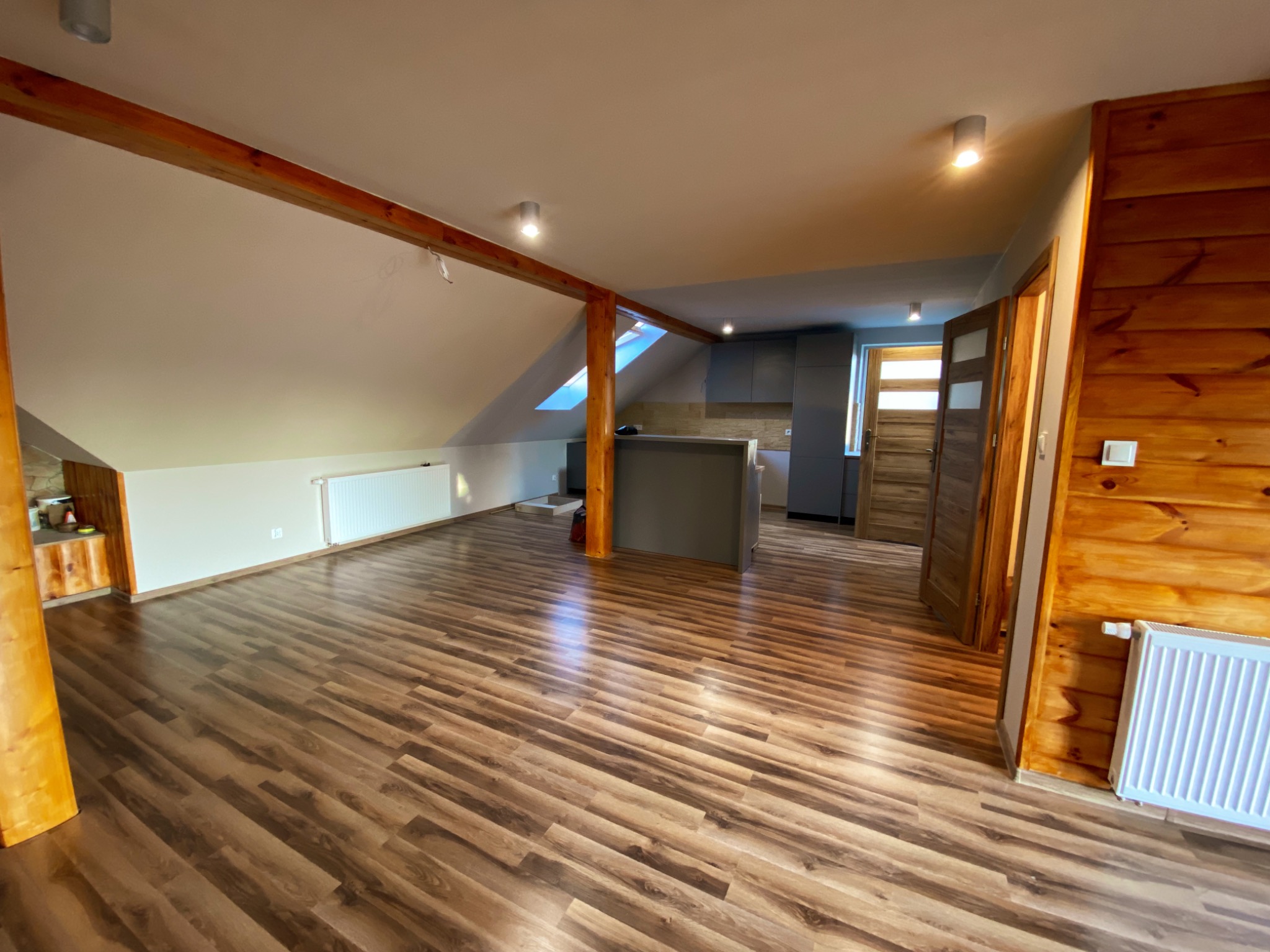 Przestronne poddasze z drewnianą konstrukcją stropu, wykończone panelami podłogowymi w odcieniach brązu, z widoczną kuchnią w tle i grzejnikiem przy drewnianej ścianie.