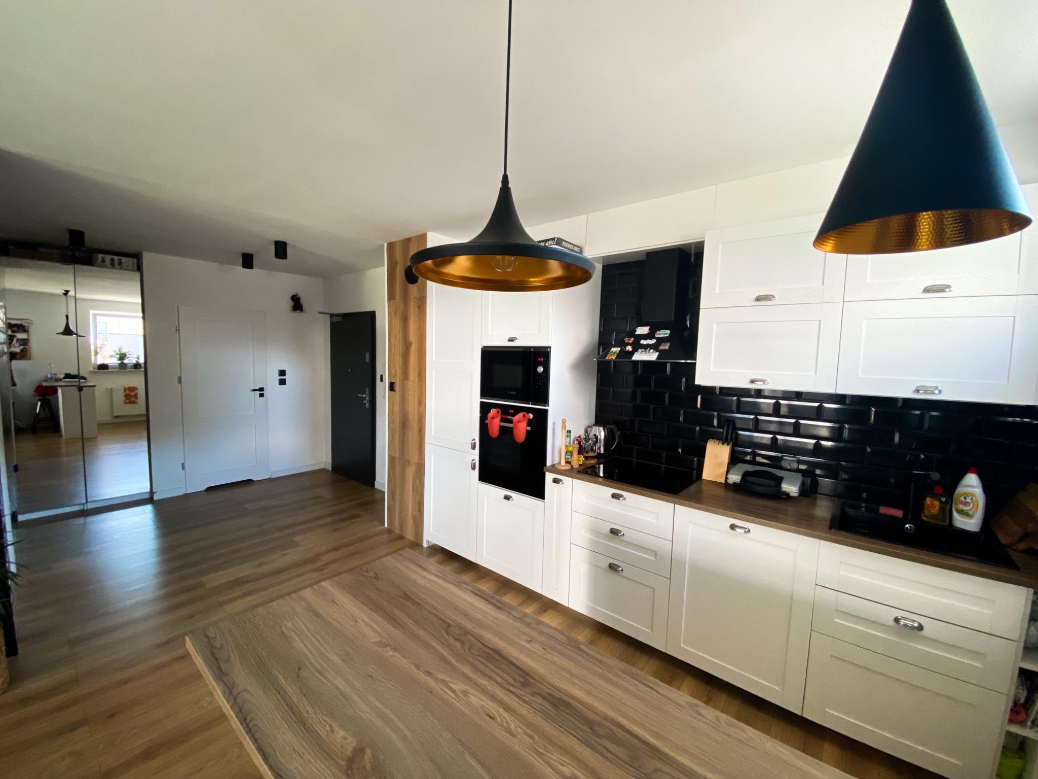 Nowoczesna kuchnia z białymi szafkami, czarną glazurą na ścianie i drewnianym stołem. Dwie wiszące lampy z czarnymi kloszami i złotym wnętrzem. Widok na przedpokój z lustrem.