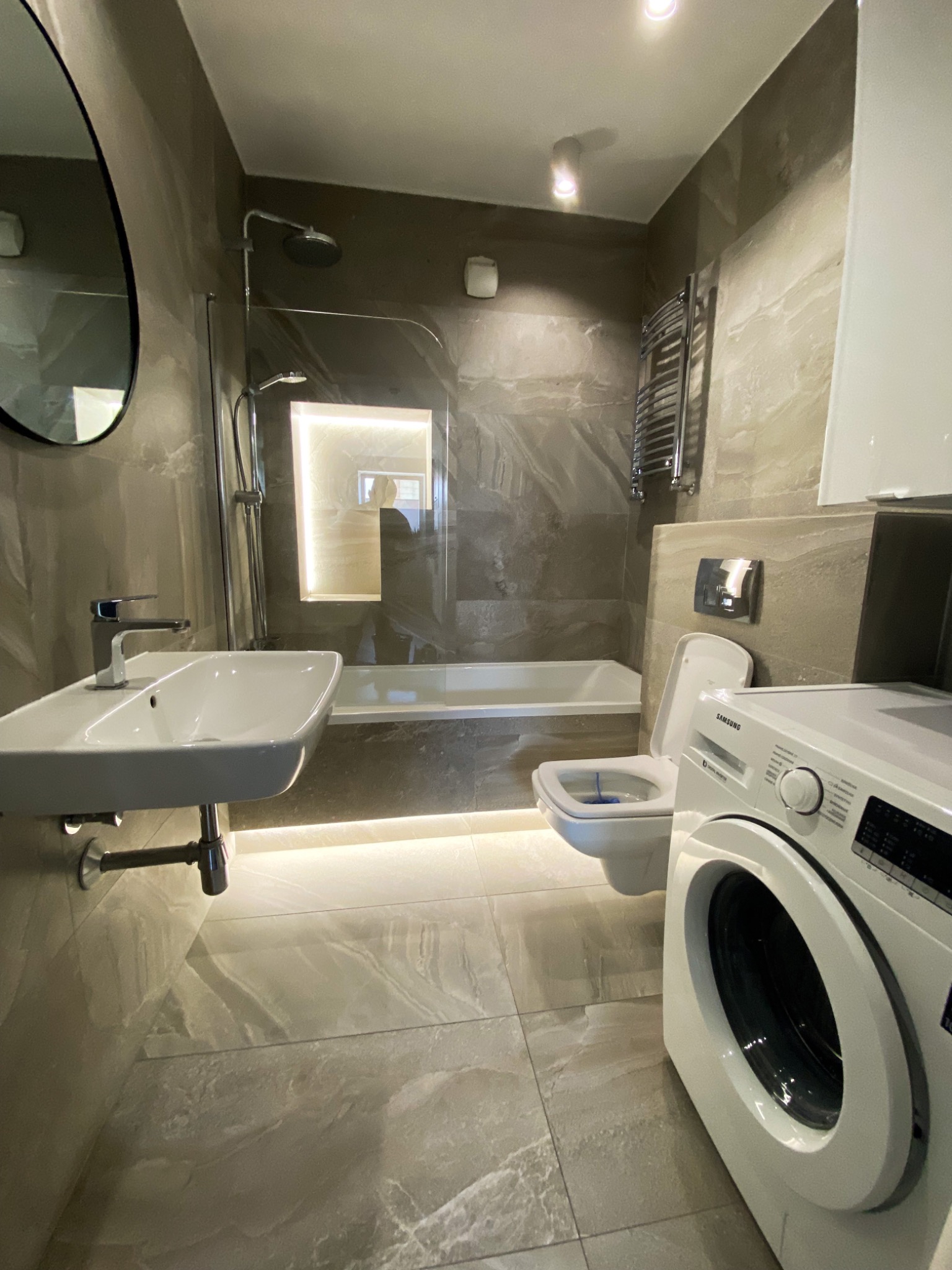 Nowoczesna łazienka z beżowymi płytkami imitującymi marmur, podświetlona listwą LED pod umywalką i wanną, z zabudowaną wnęką oświetloną w ścianie nad wanną, widoczna umywalka, toaleta, wanna...