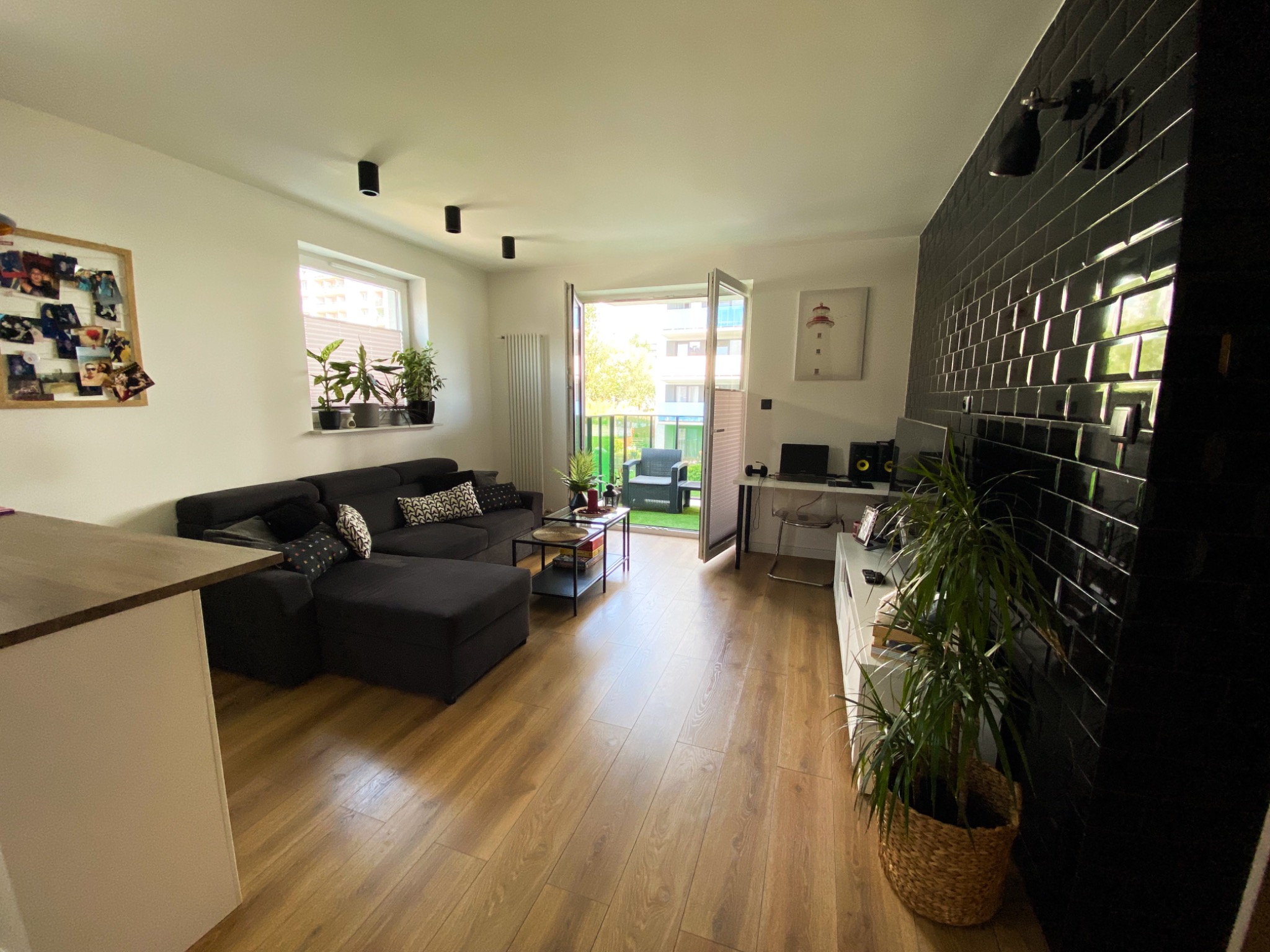 Jasne wnętrze salonu z drewnianą podłogą, czarną narożną sofą, otwartym balkonem i ścianą wyłożoną błyszczącymi, czarnymi płytkami.