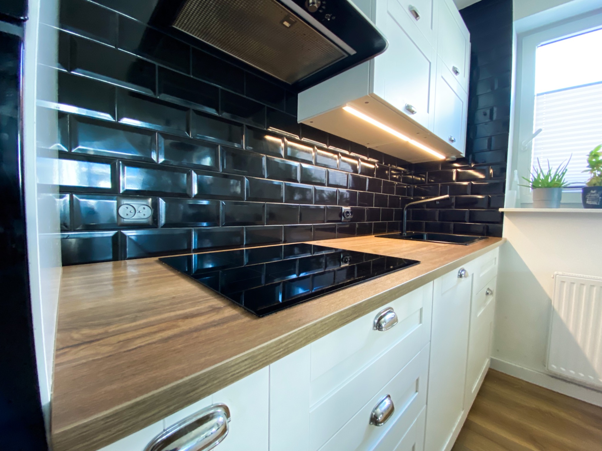 Nowoczesna kuchnia z czarnymi, błyszczącymi płytkami typu metro, drewnianym blatem, białymi szafkami z chromowanymi uchwytami i płytą indukcyjną.