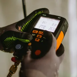 Miernik cyfrowy Testo 550 z podłączonym przewodem, trzymany w dłoniach w rękawicach roboczych, wyświetlający odczyty parametrów, prawdopodobnie podczas pomiaru czynnika chłodniczego.