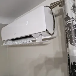 Biała klimatyzacja Gree z wyświetlaczem pokazującym 22 stopnie Celsjusza, zamontowana na szarej ścianie, obok zasłony w drzewa.