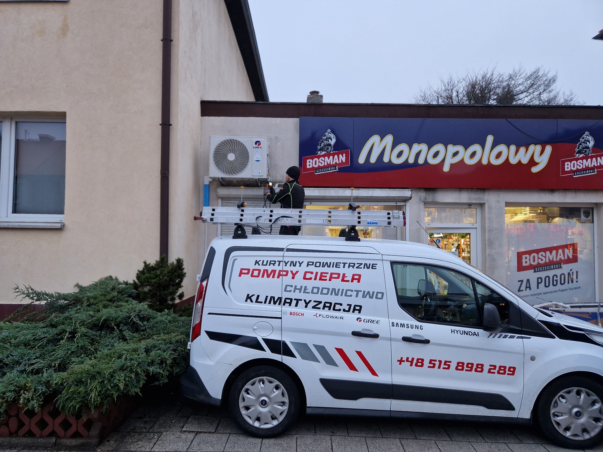 Instalator na drabinie montuje klimatyzator marki Gree na ścianie budynku z szyldem 'Monopolowy', obok zaparkowany biały samochód dostawczy z reklamą firmy instalacyjnej.