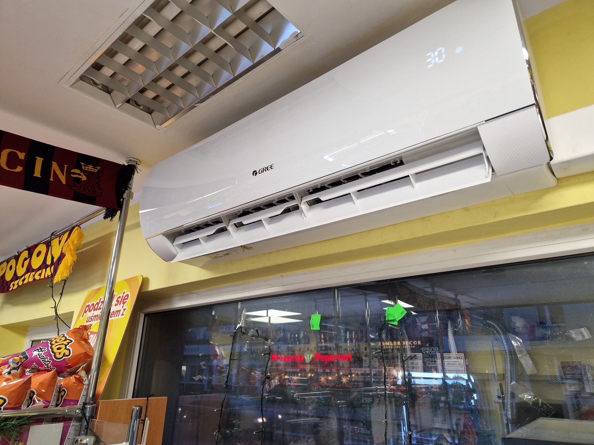 Biały klimatyzator marki Gree zamontowany w pomieszczeniu sklepowym z widocznym wyświetlaczem temperatury, na tle okna z dekoracjami i półkami z produktami.