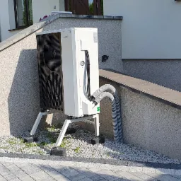 Biała pompa ciepła zamontowana na zewnątrz budynku na białym stelażu, z widocznymi szarymi przewodami, na tle elewacji i ozdobnego kamiennego podłoża. W tle częściowo widoczny biały samochód...