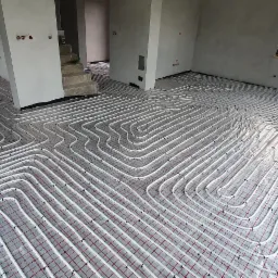 Rozłożone rury ogrzewania podłogowego na folii z siatką w surowym wnętrzu domu w trakcie budowy, widoczne okno i schody w tle.