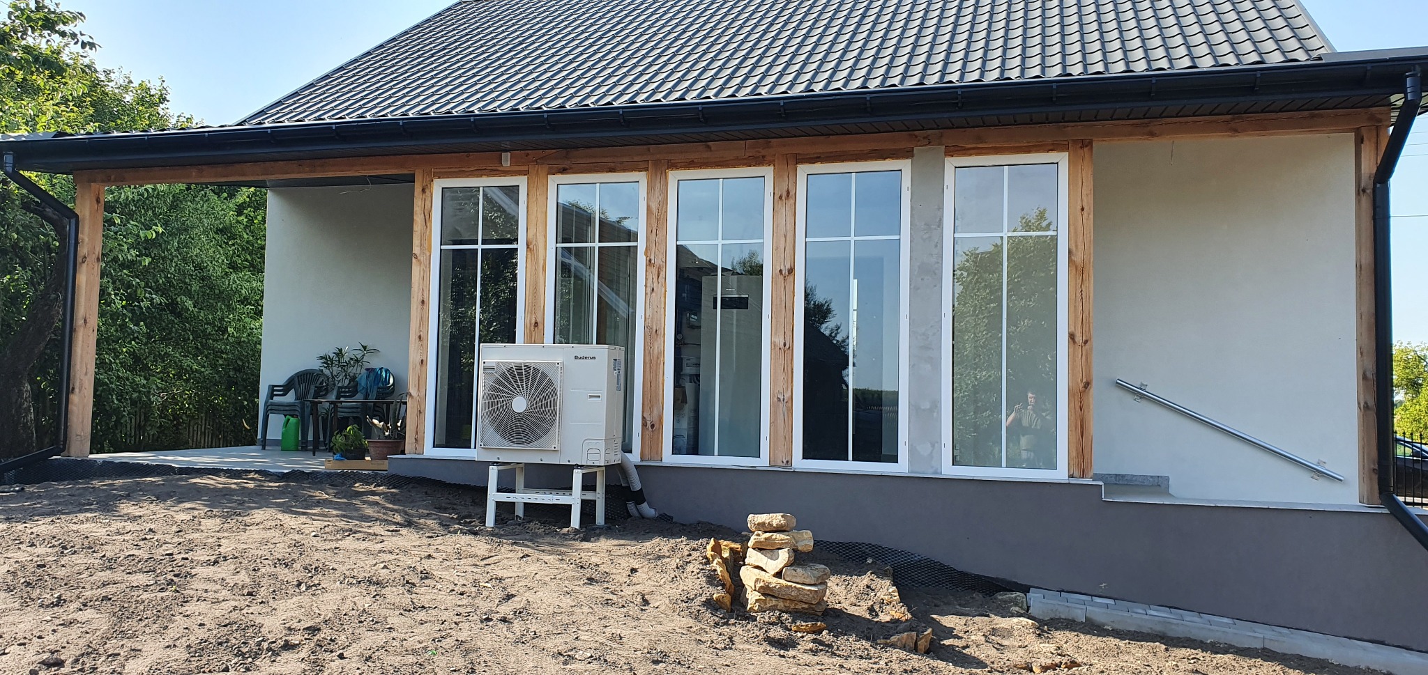 Biała pompa ciepła marki Buderus zamontowana na białym stelażu przed budynkiem z drewnianą konstrukcją werandy i dużymi oknami, widoczne ułożone kamienie dekoracyjne.