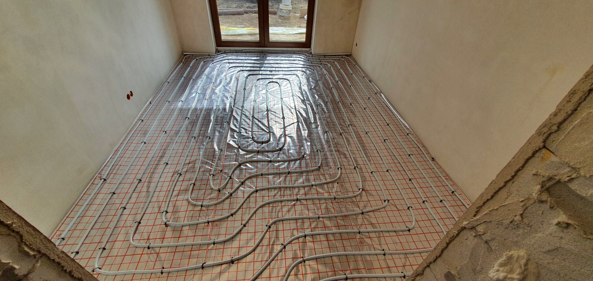 Białe rurki ogrzewania podłogowego ułożone na czerwonej siatce, przykryte folią, widok z góry, surowy stan wykończenia wnętrza z oknem w tle.