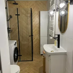 Nowoczesna łazienka z kabiną prysznicową typu walk-in, drewnopodobnymi płytkami ułożonymi w jodełkę na ścianach i podłodze, białą armaturą, czarną baterią umywalkową i lampami w metalowych kloszach.