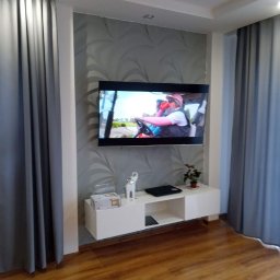 New home Krystian Ampulski - Telewizor zawieszony na ścianie z dekoracyjną tapetą, poniżej biała szafka RTV z figurką słonia i rośliną, szare zasłony po bokach, podłoga z paneli.
