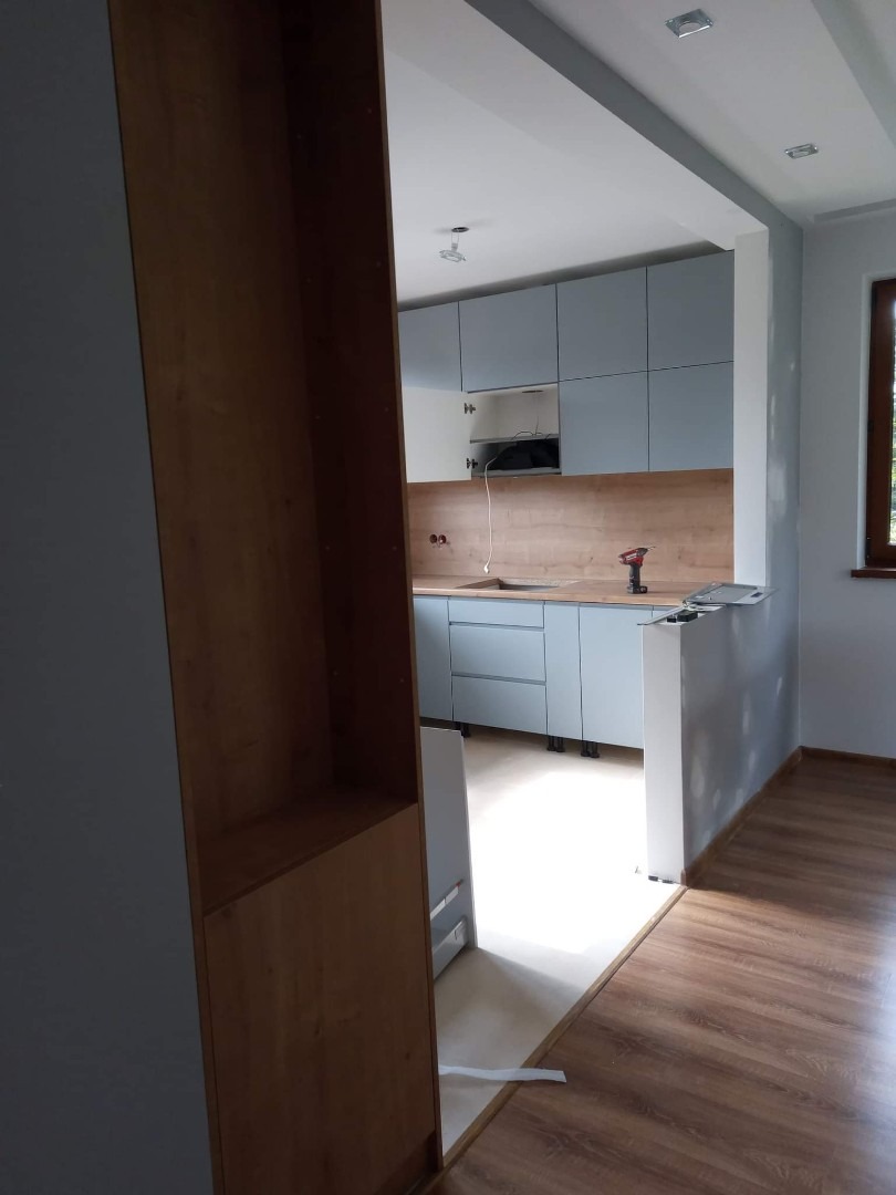 Wnętrze kuchni w trakcie montażu: widoczne szare szafki kuchenne, drewniany blat, częściowo zamontowany regał i podłoga z paneli, z wiertarką na blacie.