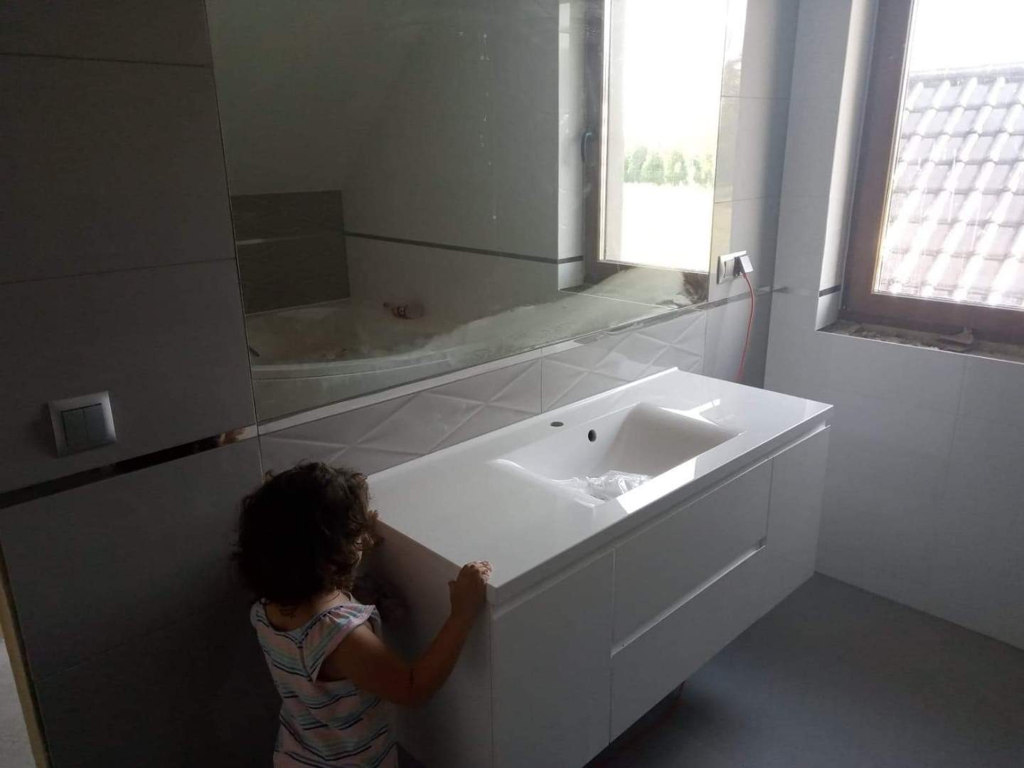 Nowoczesna łazienka z białymi płytkami, umywalką z szafką i dużym lustrem, widoczna mała dziewczynka w rogu.