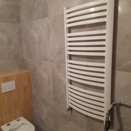 TOM-GRES - Nowoczesna łazienka z beżowymi płytkami imitującymi marmur, wisząca toaleta z drewnianą obudową i biały, designerski grzejnik drabinkowy.