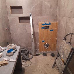 TOM-GRES - Wnętrze łazienki w trakcie wykańczania: imitacja drewnianej okładziny ukrywająca stelaż WC, szare płytki na ścianach z wnękami, poziomica oparta o ścianę, drabina i narzędzia na podłodze.
