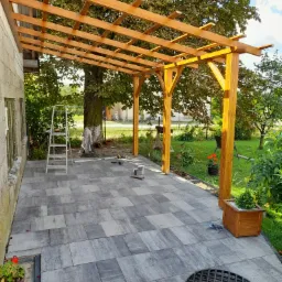 Drewniana pergola nad szarym tarasem z kostki brukowej, widoczne narzędzia i drabina, w tle zieleń ogrodu.