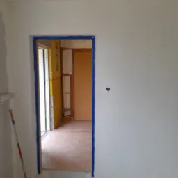 Świeżo pomalowane na biało ściany z otworem drzwiowym zabezpieczonym niebieską taśmą malarską, widoczny wałek do malowania i fragment sufitu z widocznymi łatami gipsowymi.