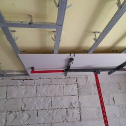 TOM-GRES - Montaż płyty na metalowym stelażu sufitowym z użyciem podpory teleskopowej na tle surowej ściany z bloków.