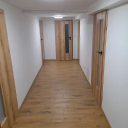 Długi korytarz z drewnopodobną podłogą, białymi ścianami i trzema drewnianymi drzwiami z przeszkleniami po obu stronach. Sufit z oświetleniem punktowym.