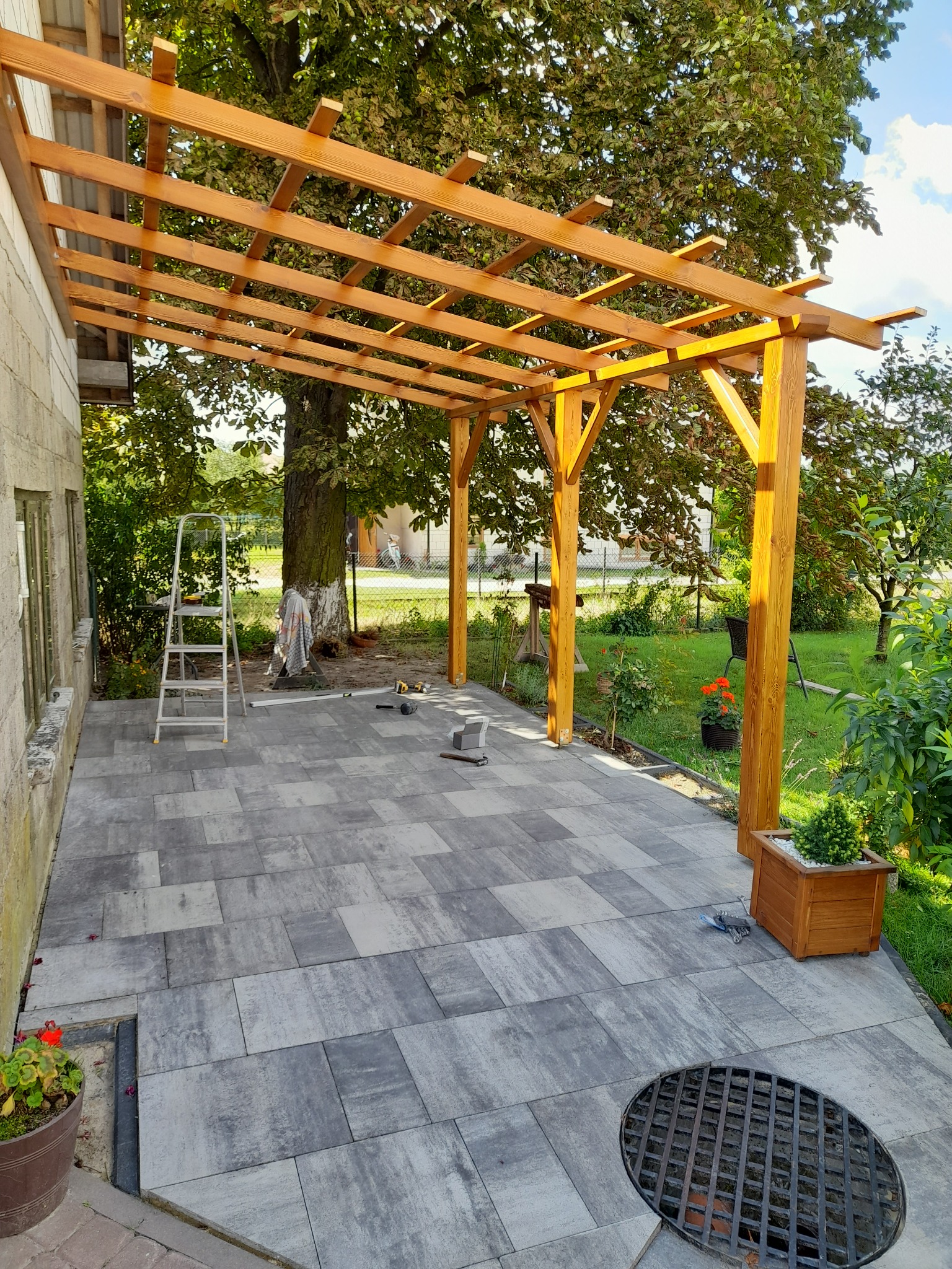 Drewniana pergola nad szarym tarasem z kostki brukowej, widoczne narzędzia i drabina, w tle zieleń ogrodu.