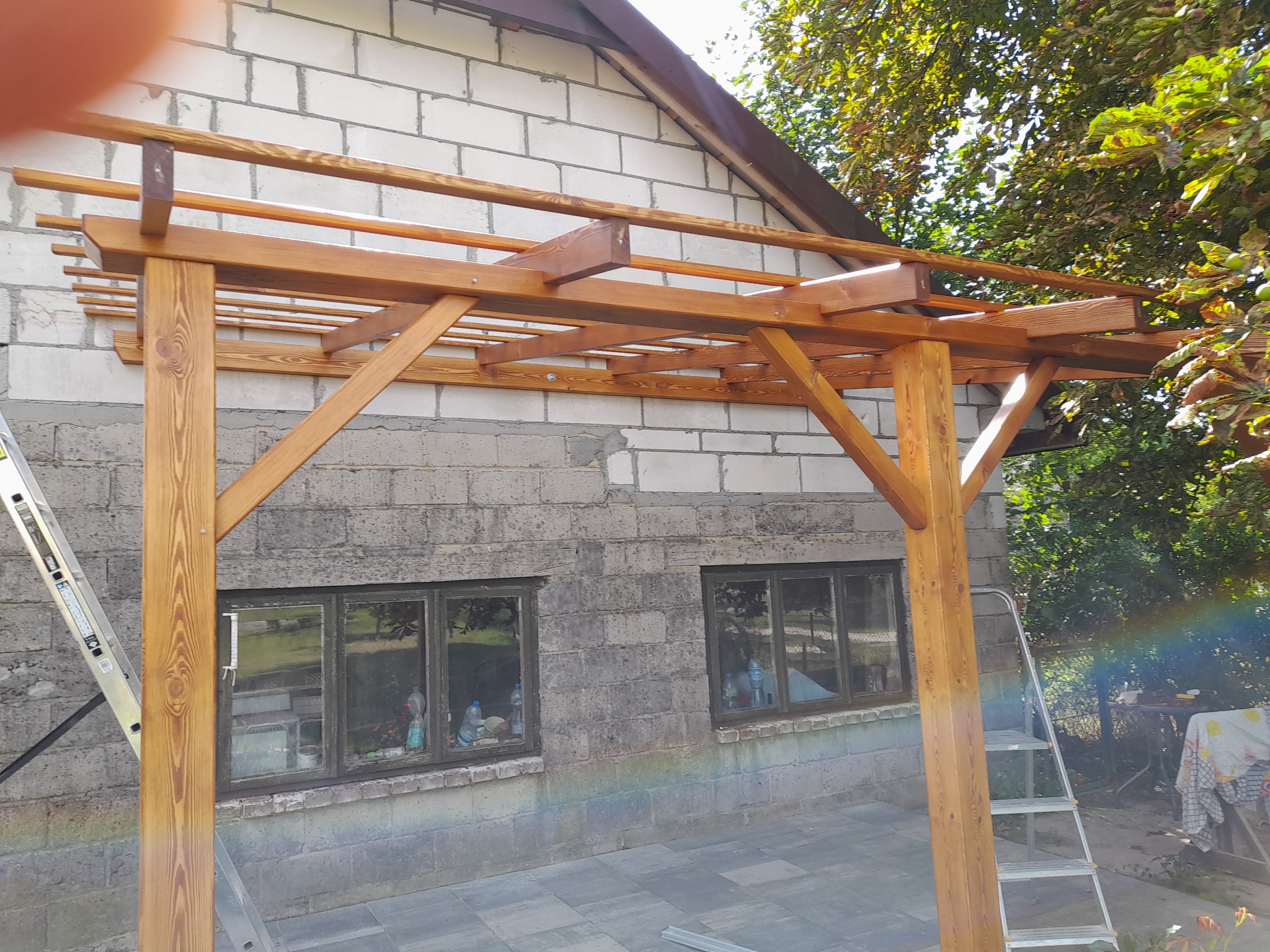 Drewniana konstrukcja z belek, prawdopodobnie zadaszenie tarasu, w trakcie montażu przy ścianie budynku z pustaków. Widoczna drabina oparta o konstrukcję i ścianę.