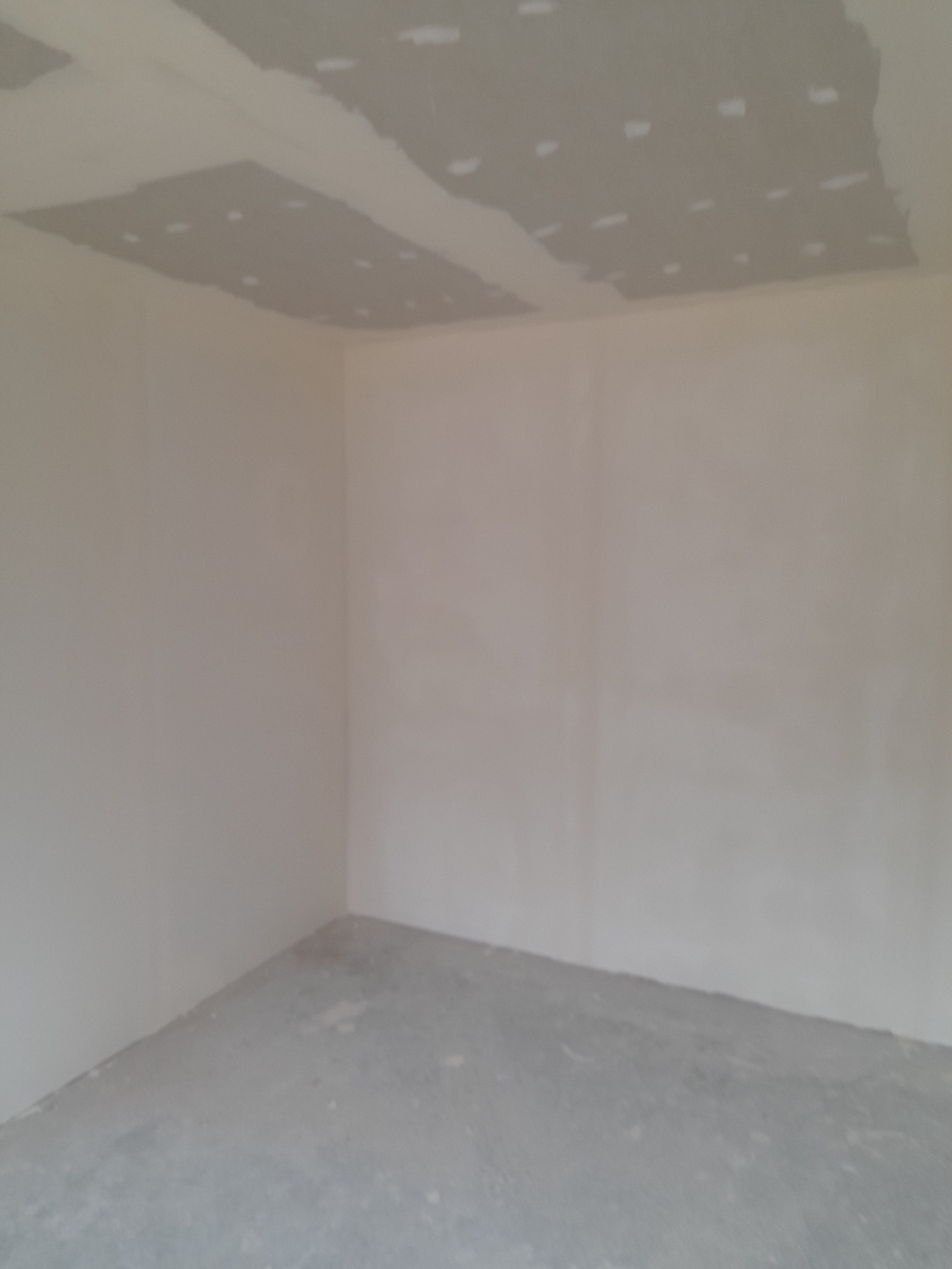 Surowe wnętrze pokoju w trakcie wykańczania: ściany pokryte warstwą szpachli, sufit częściowo z zamontowaną konstrukcją, podłoga betonowa.