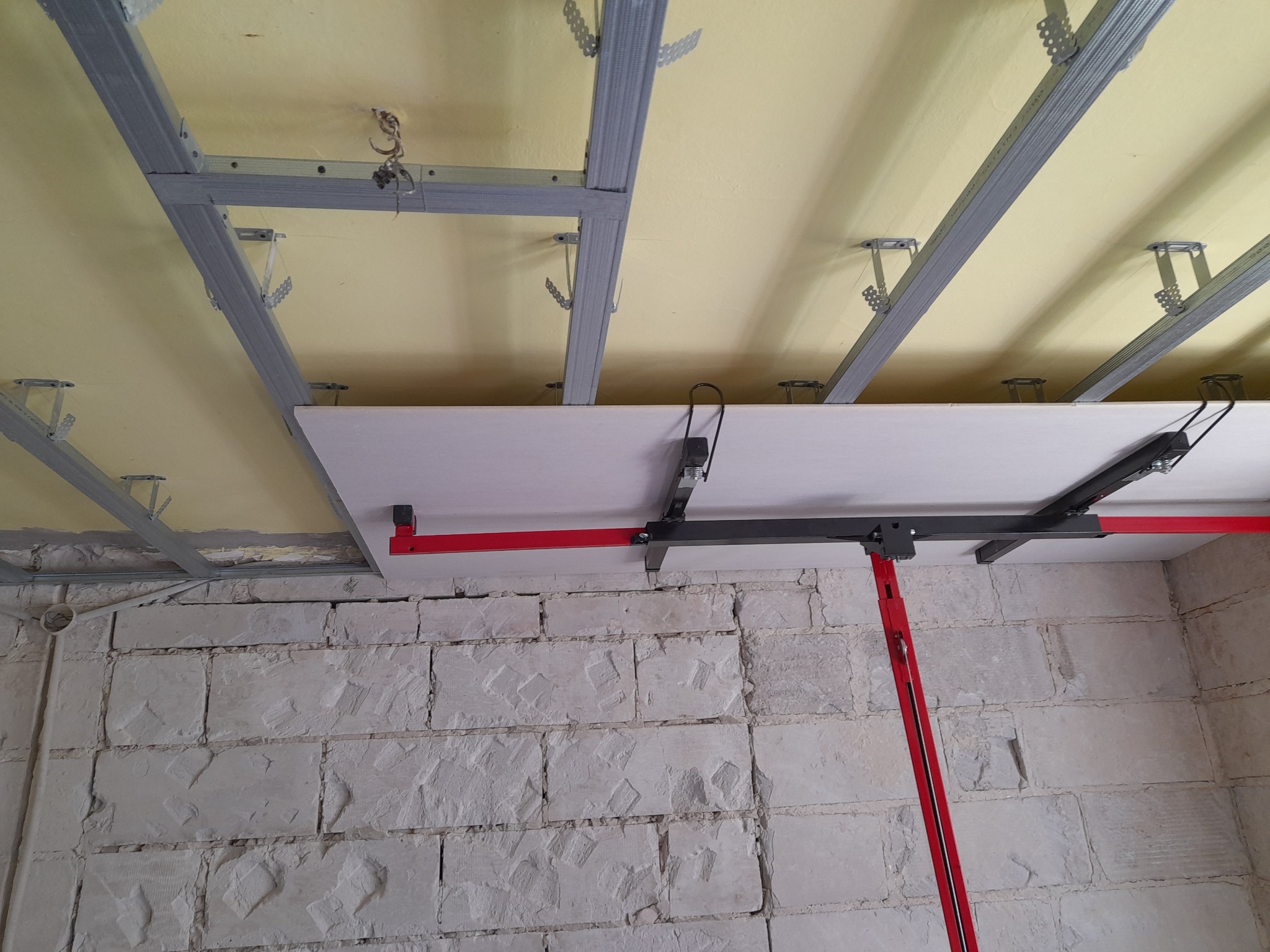 Montaż płyty na metalowym stelażu sufitowym z użyciem podpory teleskopowej na tle surowej ściany z bloków.